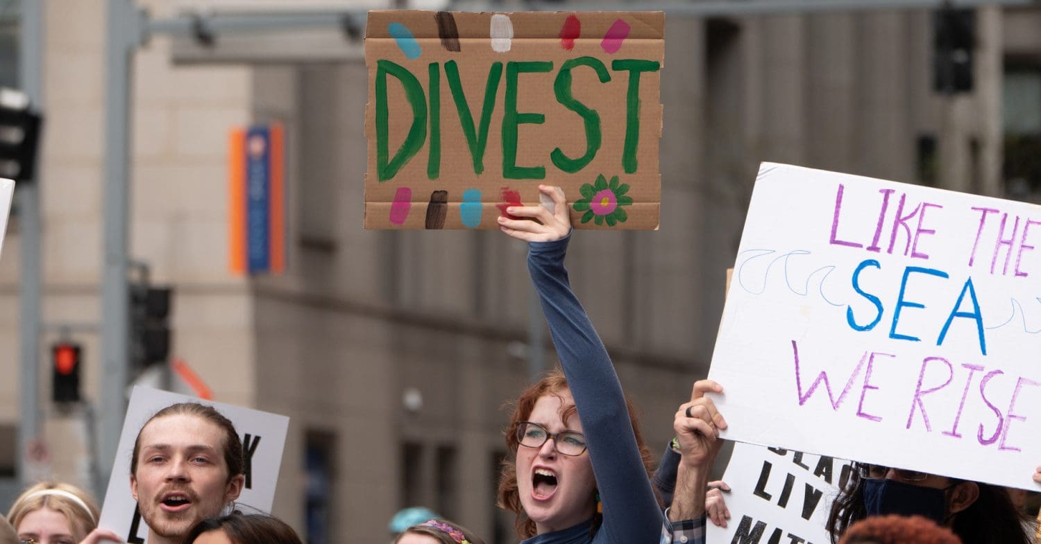 Protesters hold placards up during a climate strike. The central placard says: DIVEST.