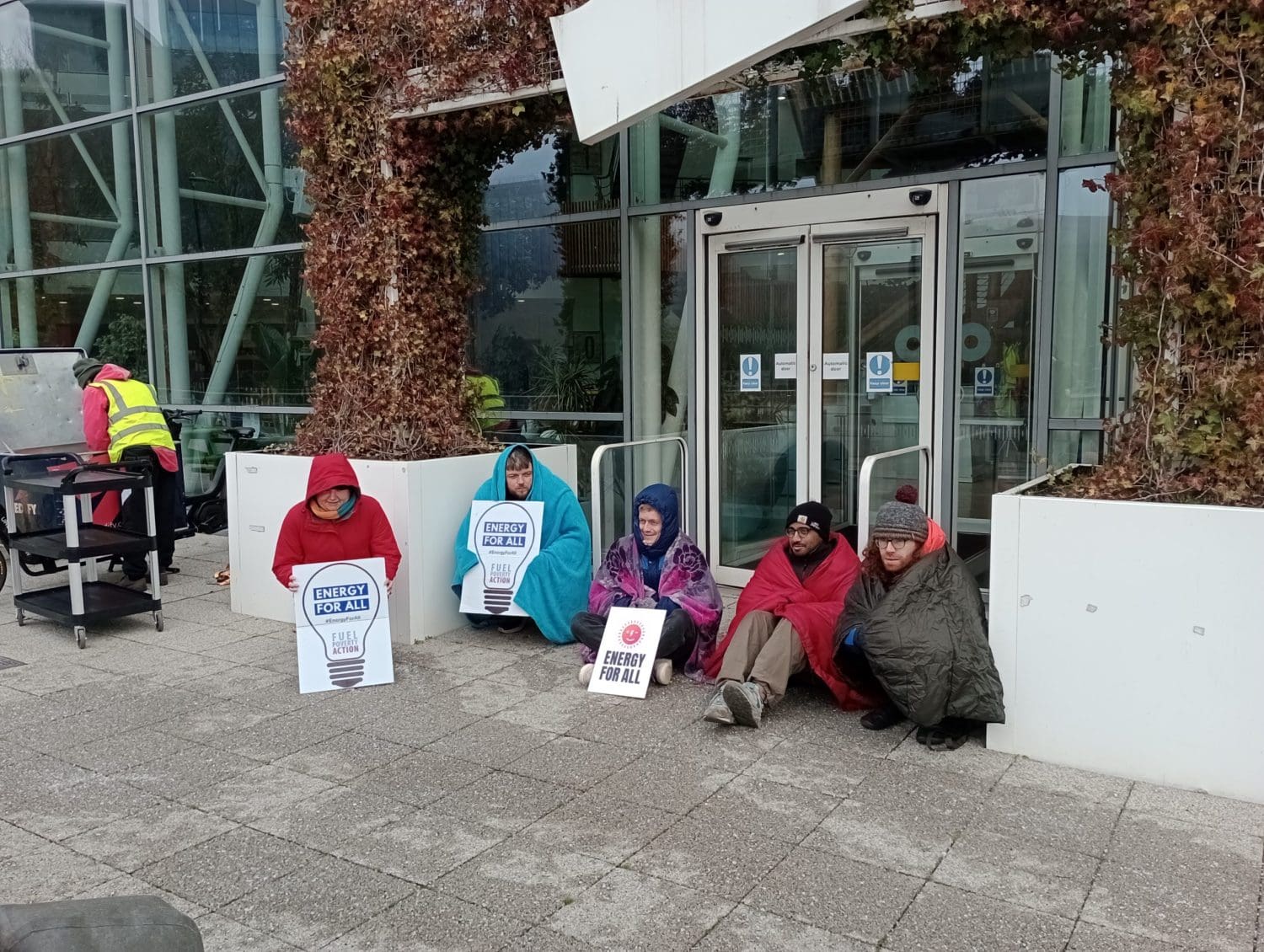 People sat on the floor outside OVO Energy's head office. They are wrapped in blankets and sleeping bags and have signs that read "energy for all" 