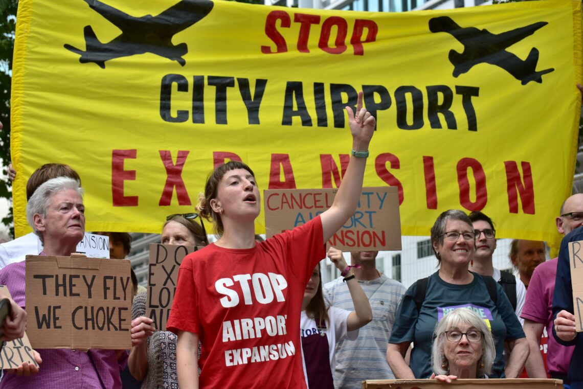 Protesters gather with banners and placards which read: "Stop City Airport Expansion", "Revoke the decision", "They fly, we choke", and "Ban expansion, not protest". 