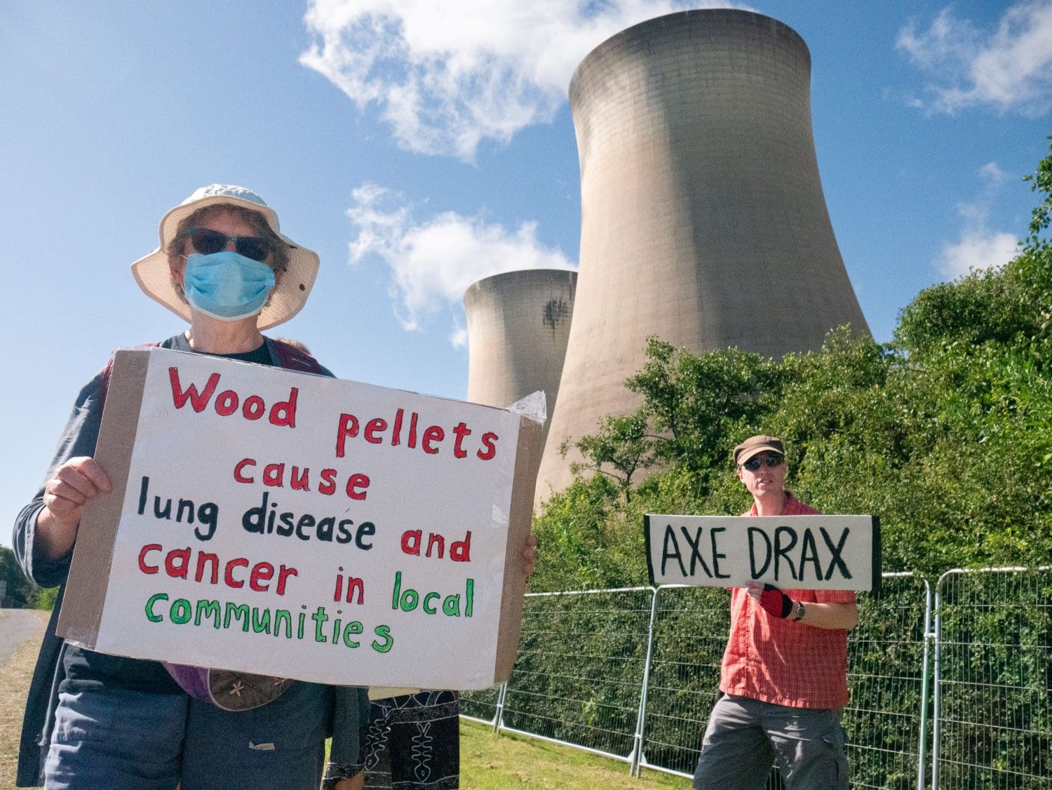 Drax towers loom over two protesters holding placards reading: "Wood pellets cause lung disease and cancer in local communities." and "Axe Drax". 