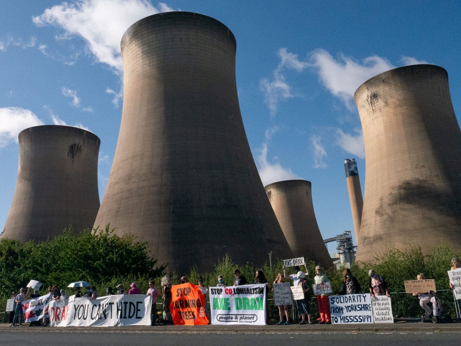 Climate protesters form a line with banners and placards. Drax flue stacks loom behind. Banners and placards read from left to right: "Count Drax-ula", "Solidarity from Yorkshire to Mississippi", "Drax: You can't hide", "Stop burning trees", "Stop CO2lonialism - Axe Drax - People & Planet Sheffield". 