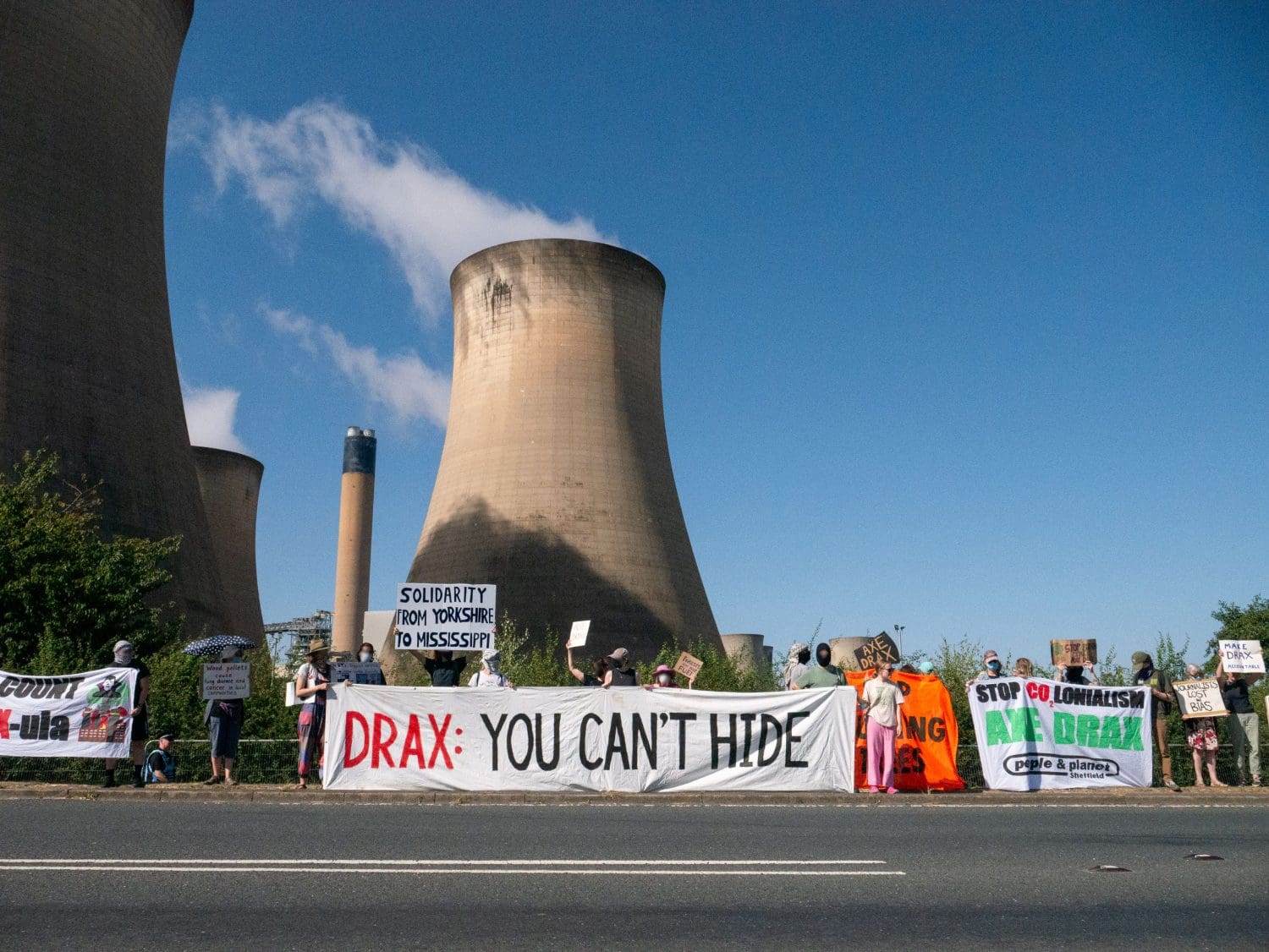 Climate protesters form a line with banners and placards. Drax flue stacks loom behind. Banners and placards read from left to right: "Count Drax-ula", "Solidarity from Yorkshire to Mississippi", "Drax: You can't hide", "Stop burning trees", "Stop CO2lonialism - Axe Drax - People & Planet Sheffield". 