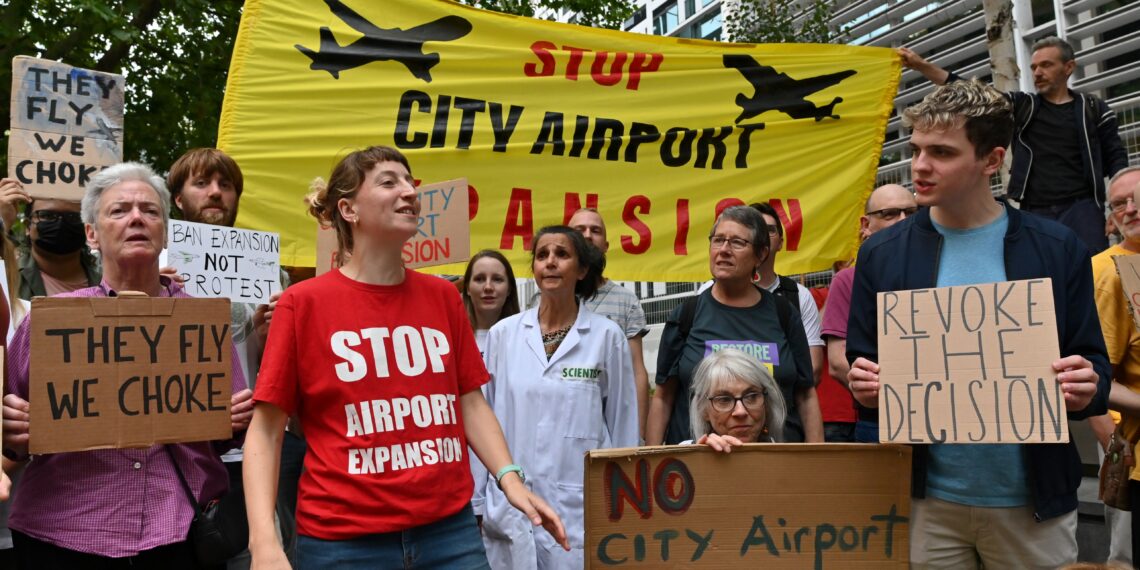 Protesters gather with banners and placards which read: "Stop City Airport Expansion", "Revoke the decision", "They fly, we choke", and "Ban expansion, not protest". 