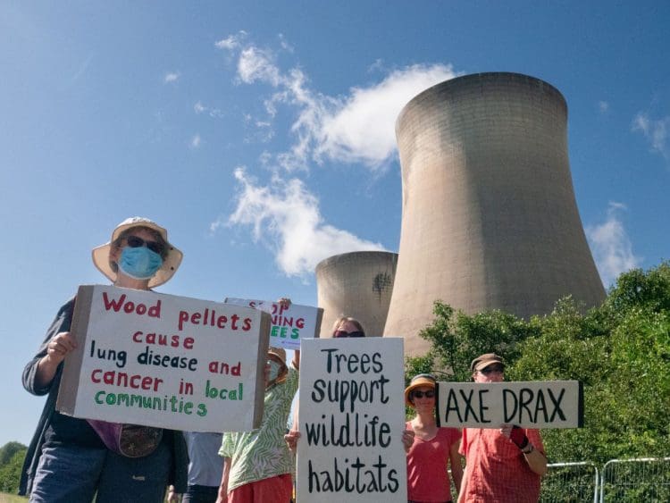 Climate protesters stand under enormous gas flue stacks at Drax power station near Selby, Yorkshire. They hold placards calling out the company's complicity. They read: "Wood pellets cause lung disease and cancer in local communities", "Trees support wildlife habitats" and "Axe Drax".