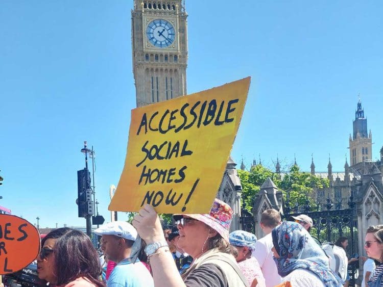 Paula Peters holding a yellow placard that reads in black writing "accessible social homes now" with Big Ben in the background. It is a sunny day at a protest over accessible housing