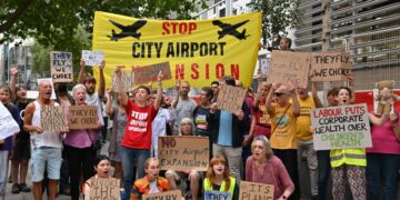 Protesters gather with banners and placards which read: "Stop City Airport Expansion", "Revoke the decision", "They fly, we choke", and "Ban expansion, not protest" London City Airport Labour