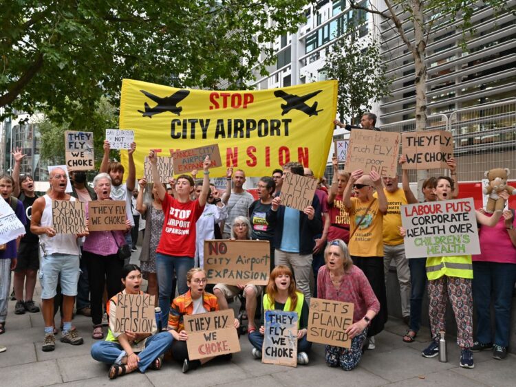 Protesters gather with banners and placards which read: "Stop City Airport Expansion", "Revoke the decision", "They fly, we choke", and "Ban expansion, not protest" London City Airport Labour