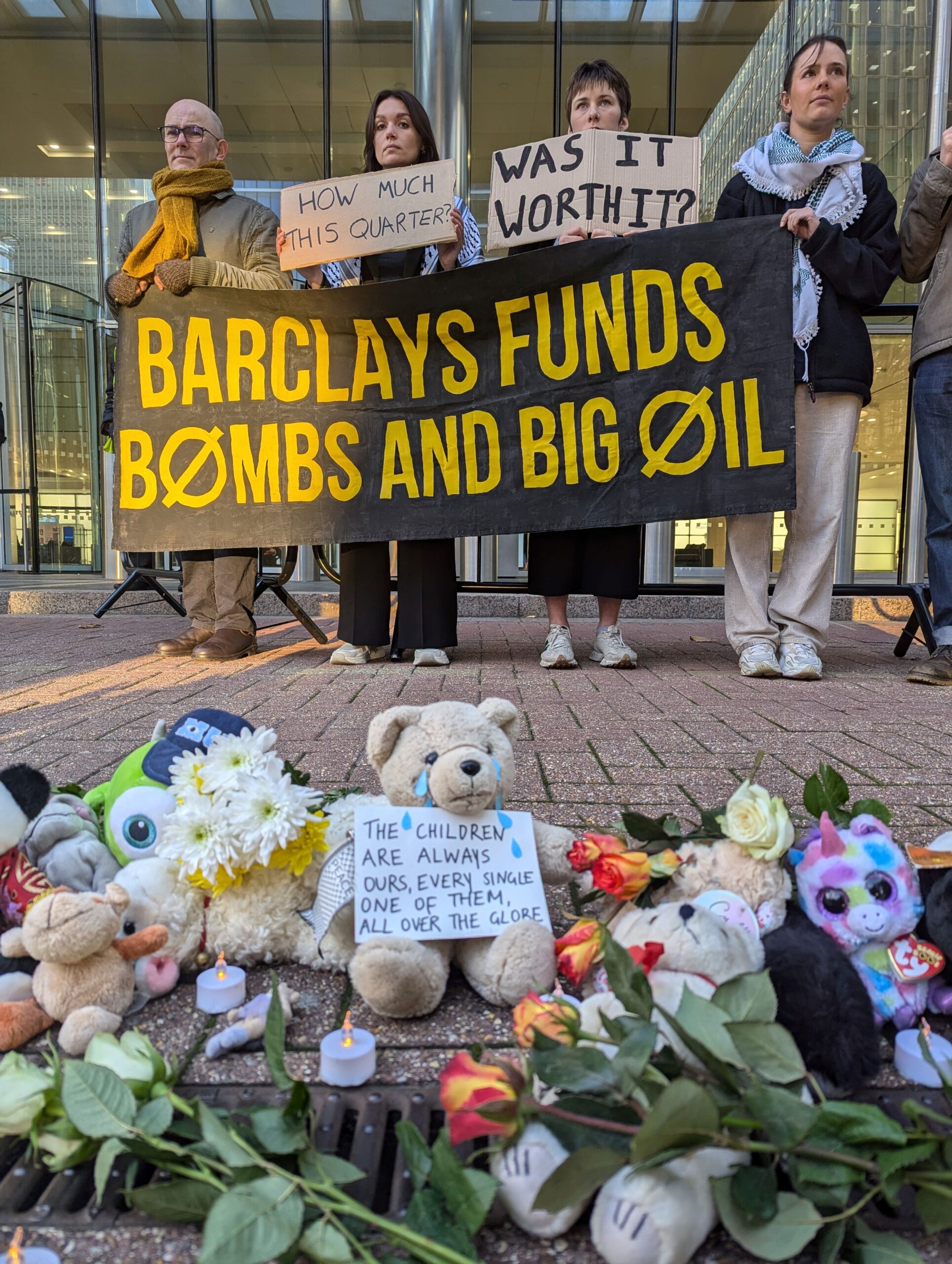 Protesters stand in a line behind a collection of soft toys and flowers laid out on the ground. They hold a banner that reads: "Barclays funds bombs and big oil" and placards that say: "How was this quarter?" and "Was it worth it?"