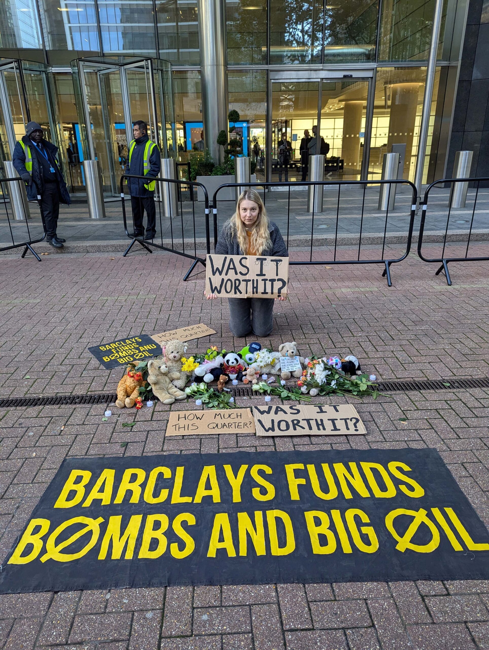 Protester holding a placard that reads "Was it worth it?" kneels behind soft toys and other placards, and a banner that reads: "Barclays funds bombs and big oil".