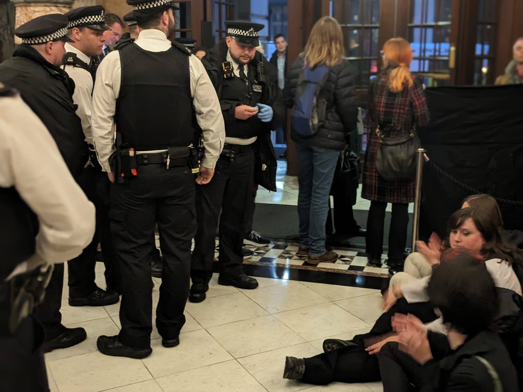 Group of upwards of six cops stand watching a small group of protesters sit in the foyer. 