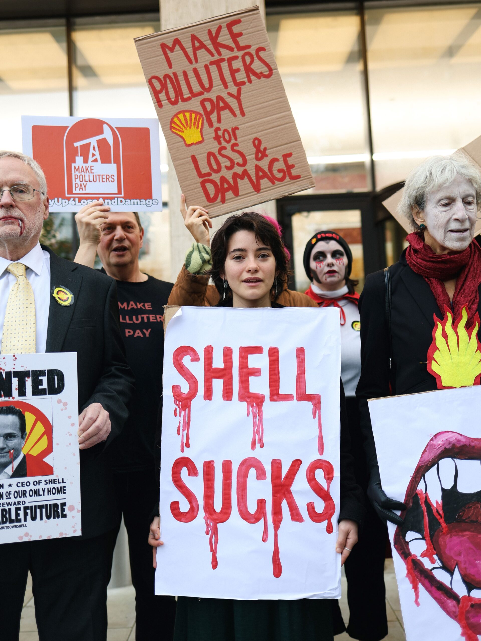 Activists dressed as ghouls and vampires hold placards reading "Shell sucks" and "Make polluters pay for loss & damage", a burning Shell logo, and vampire teeth dripping in blood. 