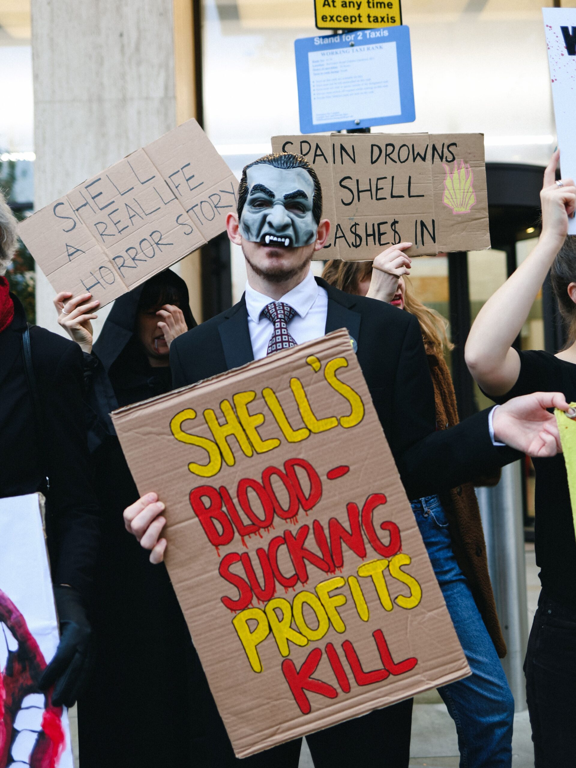 Black-robed activist holds a sign reading "Shell - a real life horror story", while another dressed as Dracula holds one that states "Shell's bloodsucking profits kill". Other activists hold up signs beside them reading "Spain drowns, Shell ca$he$ in". Shell profits