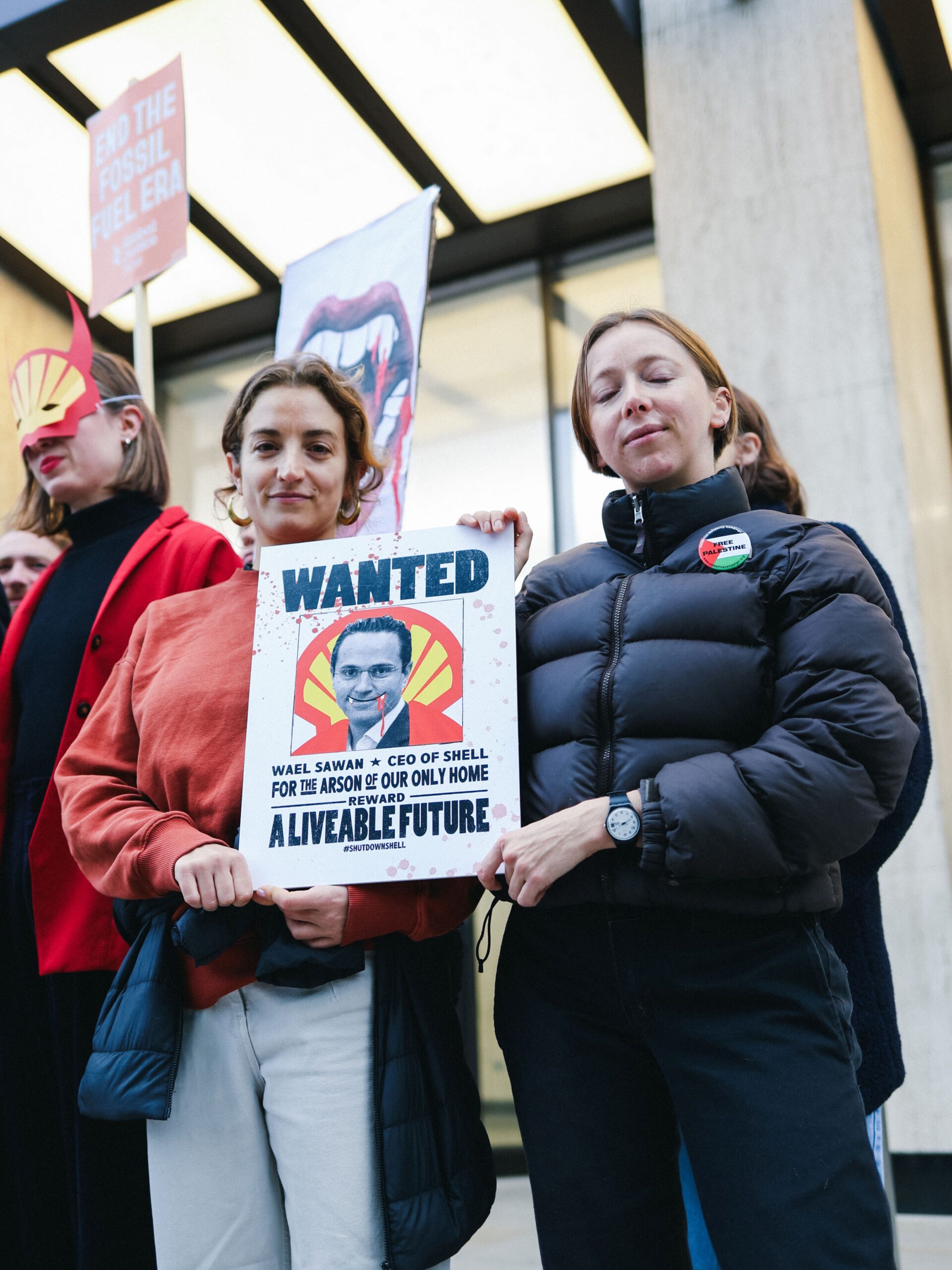 Activist in Shell logo mask with devil horns. Two other activists holding a placard that reads: "Wanted: Wael Sawan * CEO of Shell - for the arson of our only home. Reward: A liveable future. #Shutdownshell