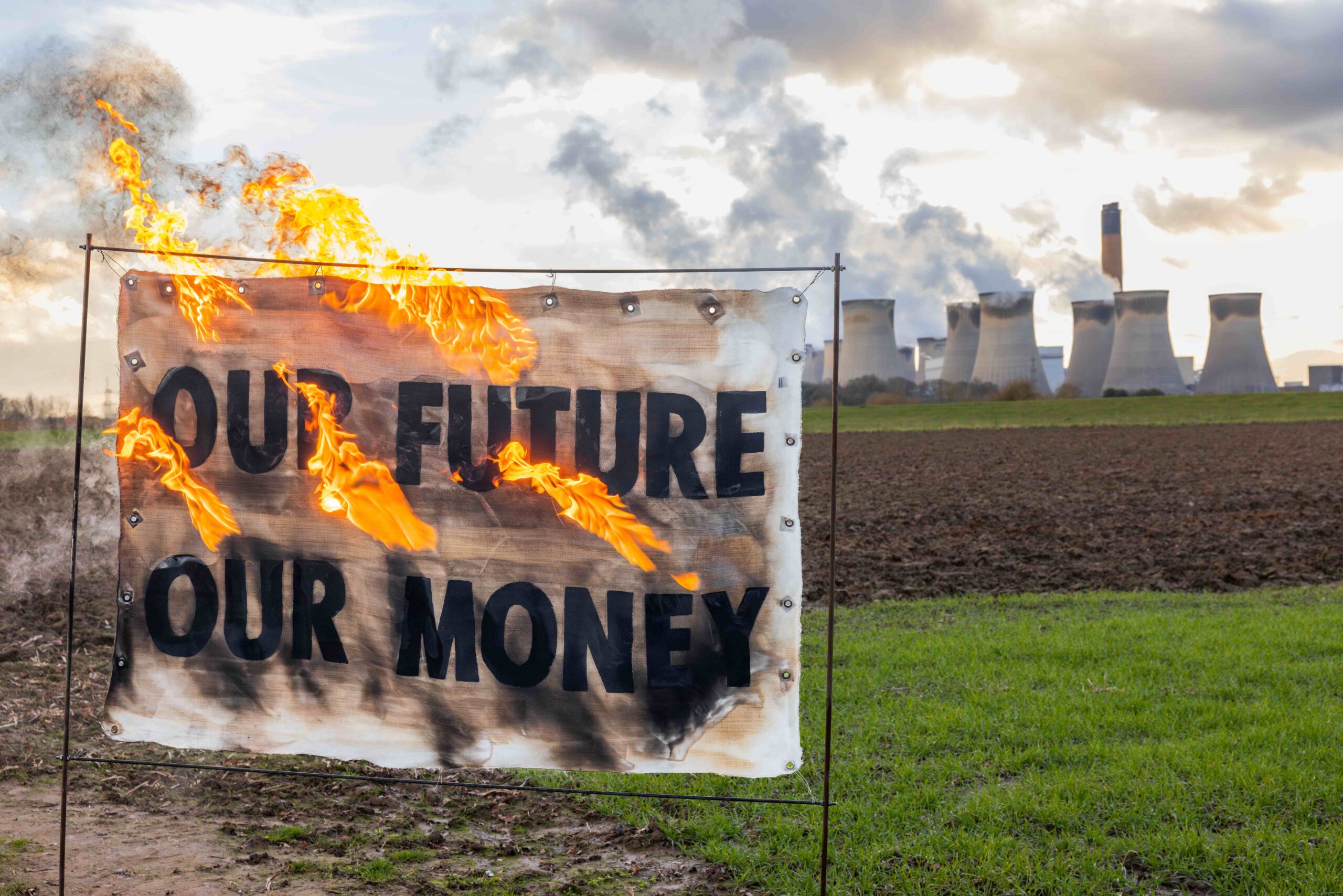 Banner on fire in front of Drax power station that reads: "Our future - our money", with smoke rising in the background from the smokestacks.