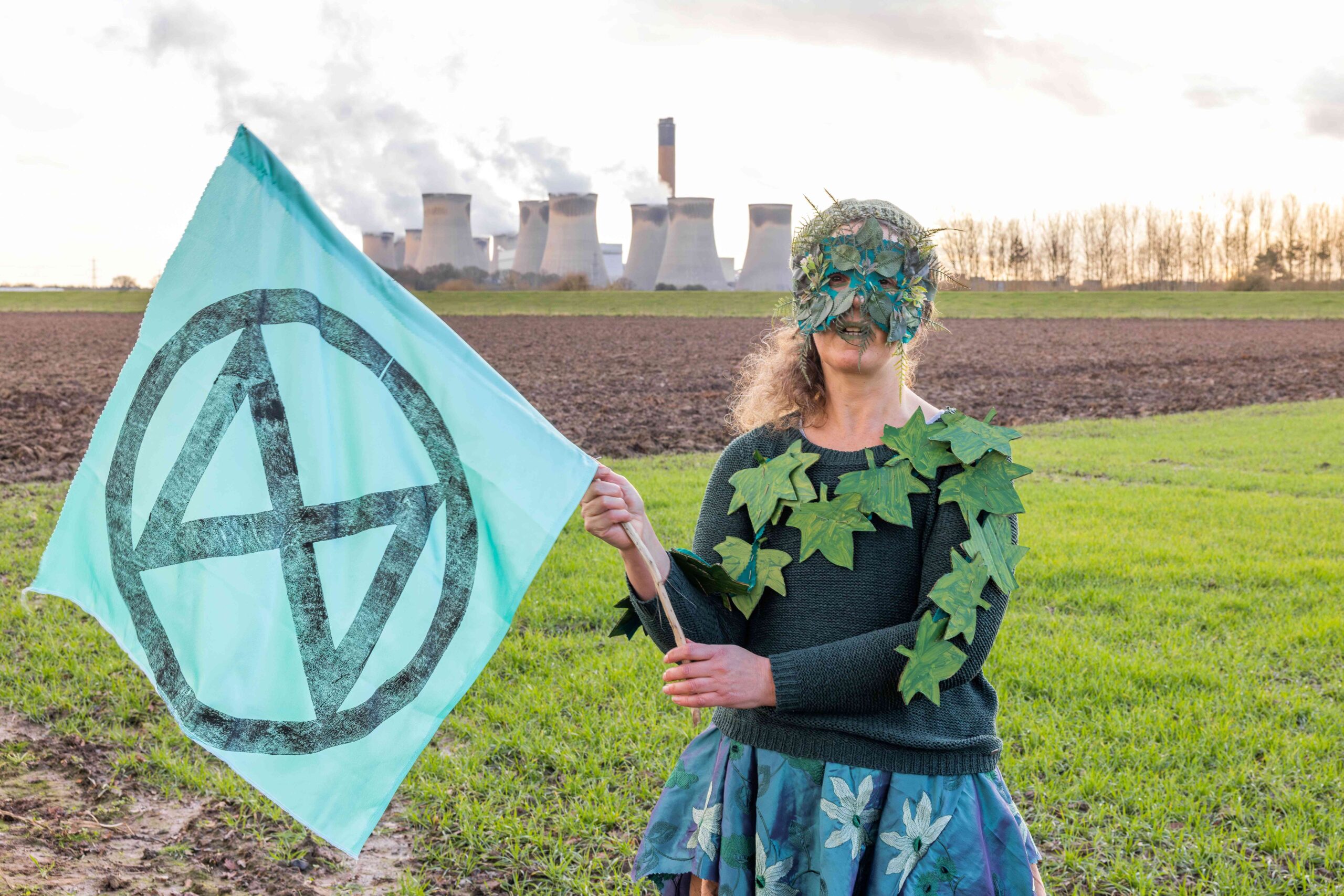 Protester with a leafy eye-mask and leaves across her top waves an Extinction Rebellion flag in front on Drax's power station smokestacks. 