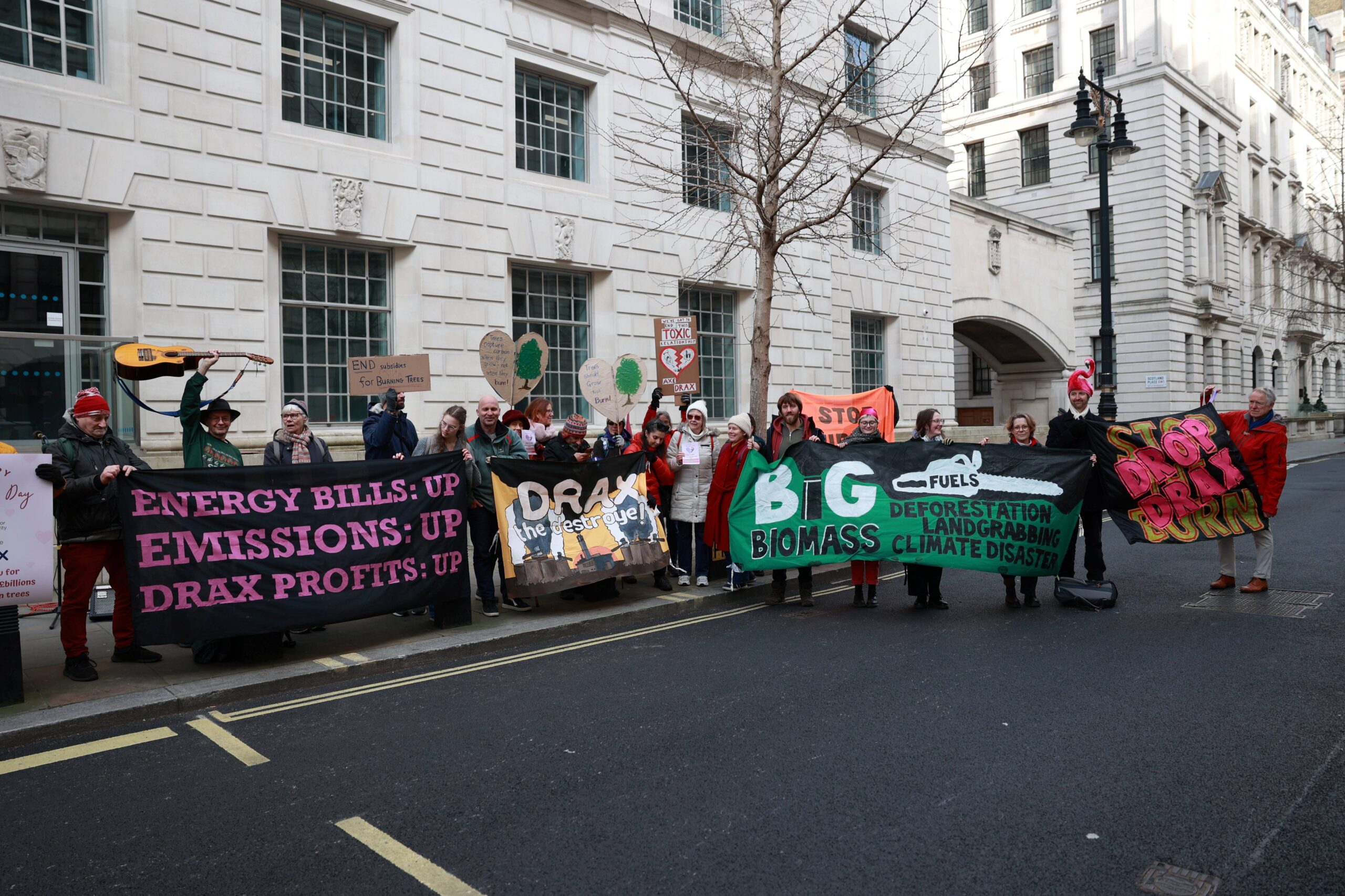 Protesters stand together with banners. Left banner black with pink text reads: "Energy bills: up, Emissions: up, Drax profits: up. Banner in the centre reads: "Drax the destroyer". Next banner over reads: "Big biomass fuels deforestation, landgrabbing, climate disaster. Banner on the right reads: Drop Drax. 