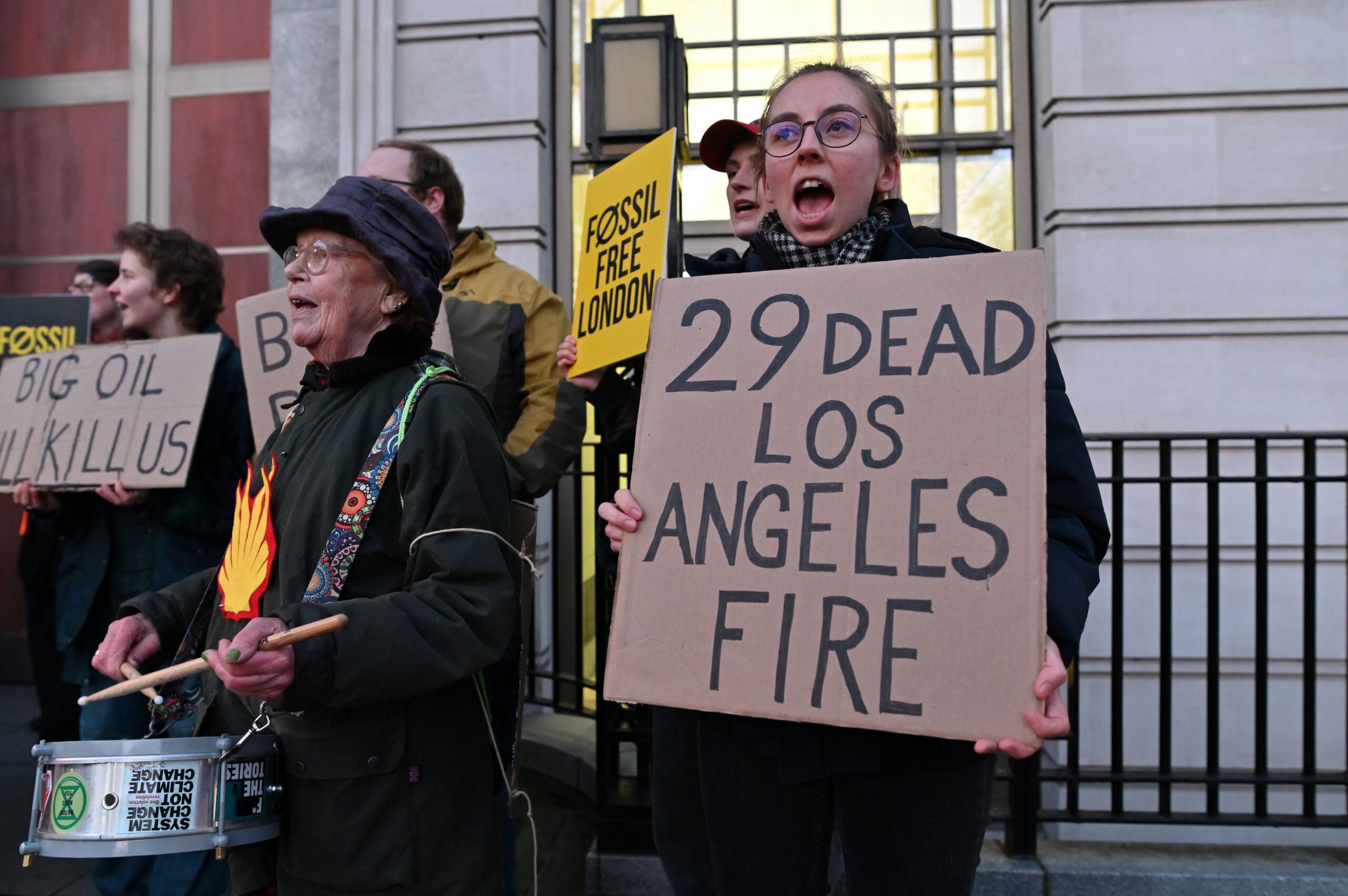 Protester holds a placard that reads "29 dead Los Angeles fire" while another drums beside her. 