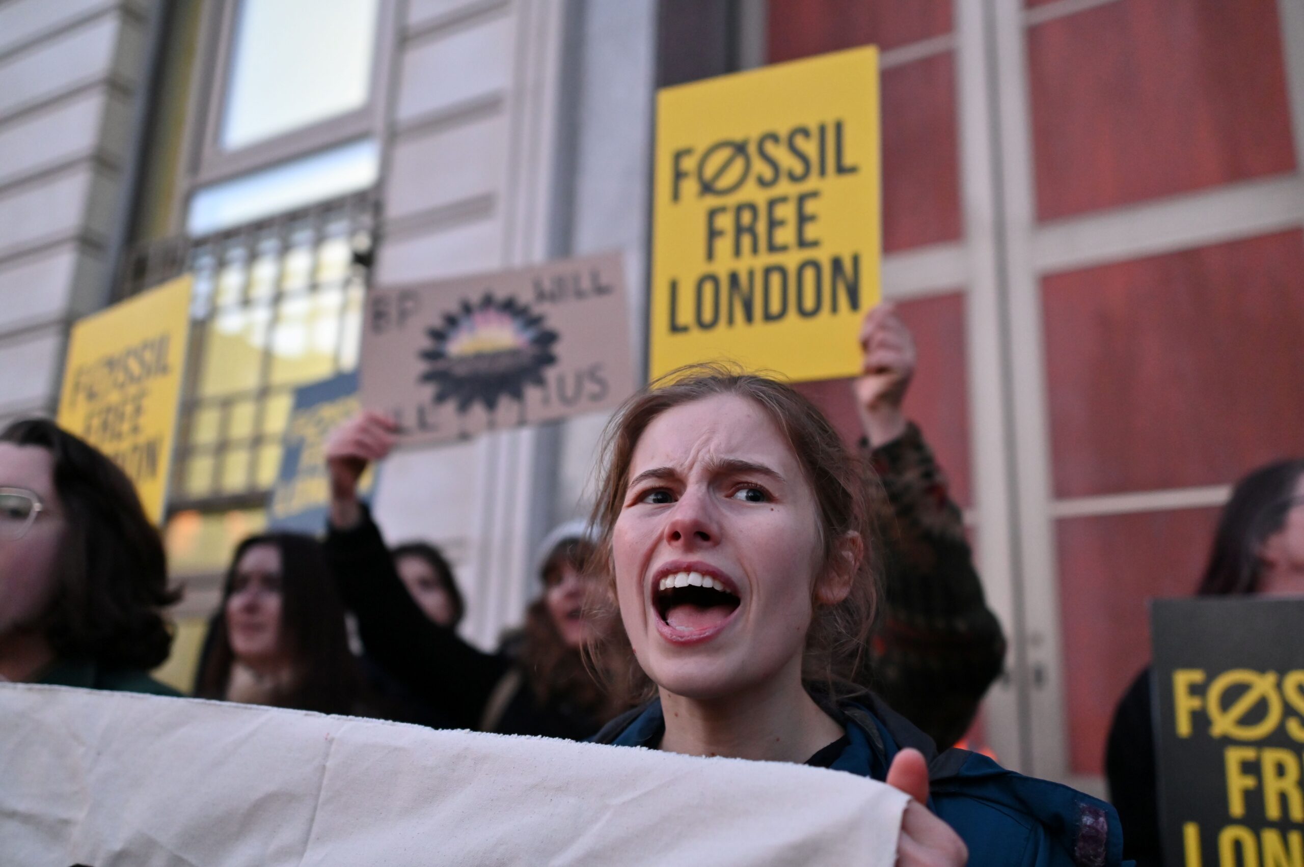 Female protester holding the group's main banner in focus, shouting. Other raise "BP will kill us" and "Fossil Free London" placards behind. 