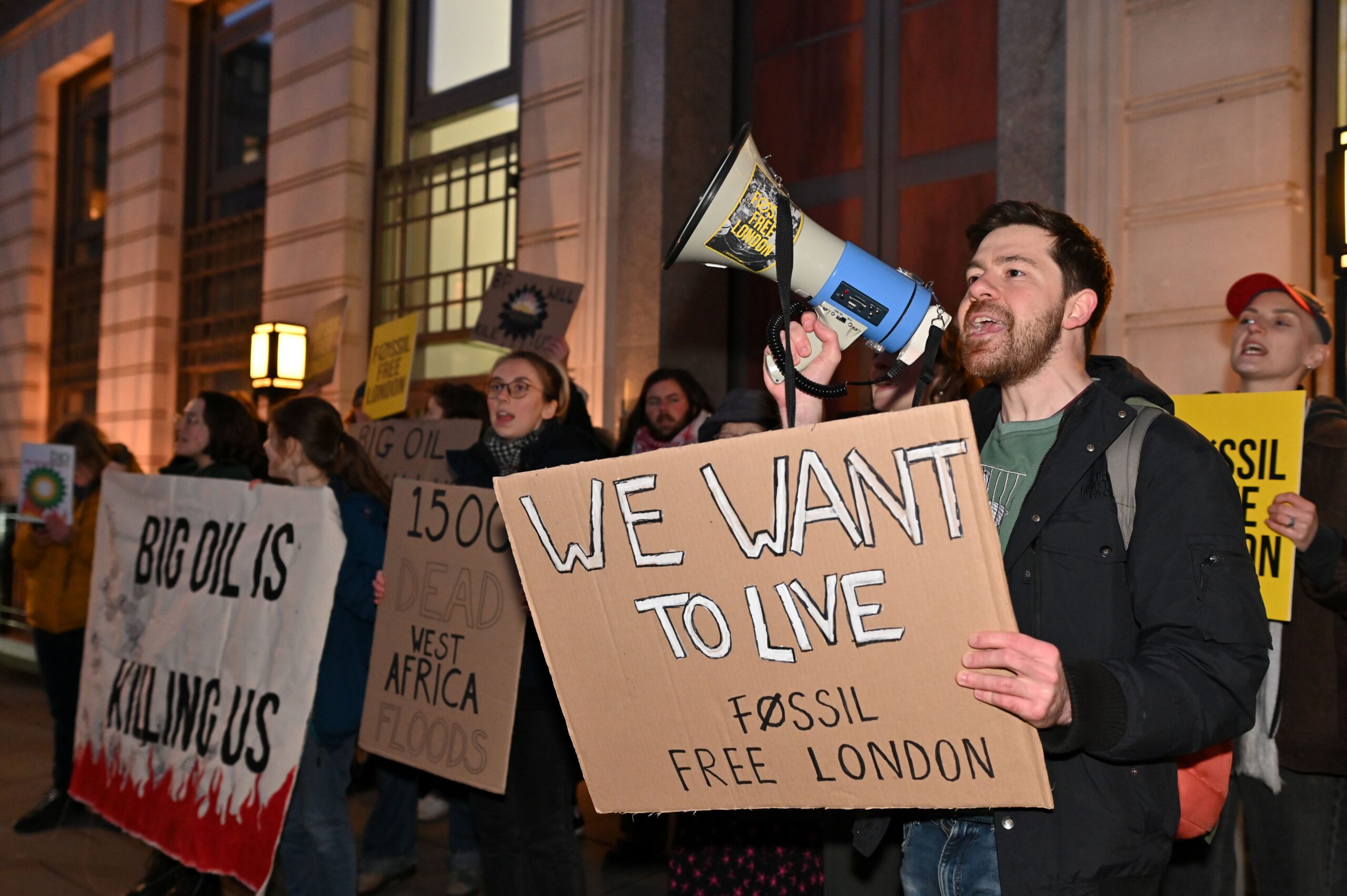 Side on view of protesters in a crowd outside the HQ with various banners and placards. To the left a banner reads "Big oil is killing us" with red flames. In the centre, a protester holds a placard reading: "1500 dead West Africa floods". On the right, a protester holds a megaphone and a placard that reads: "We want to live - Fossil Free London".