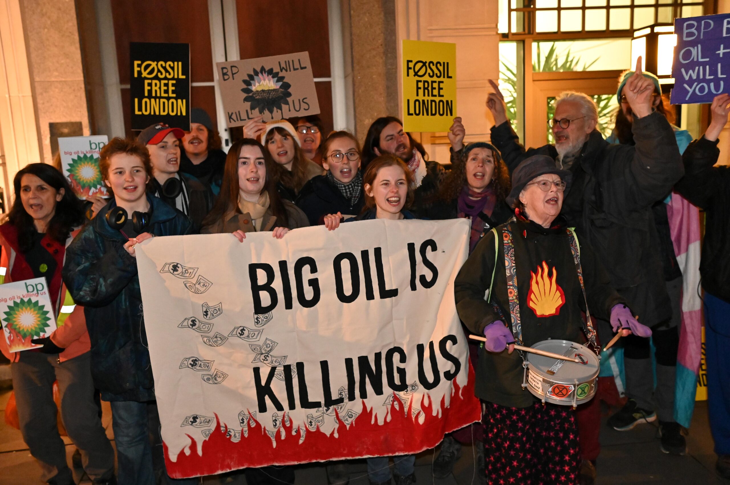 Around 15 protesters gathered in a group outside BP's HQ. Some carry placards with BP's logo on fire, while others raise Fossil Free London signs above their heads. A campaigner holds a placard with BP's sunflower logo transformed into a burning sunset and oil with the words: "BP will kill us". At the front, protesters hold a banner with dollar bills floating down to red flames and big bold text that reads: "Big oil is killing us.". A person drums at the front of the group BP renewables