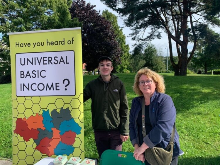 Louis Strappazzon and Alison Hawdale from the UBI Lab Network stand behind a table with campaign materials. An upright banner beside them reads: "Have you heard of a Universal Basic Income?"