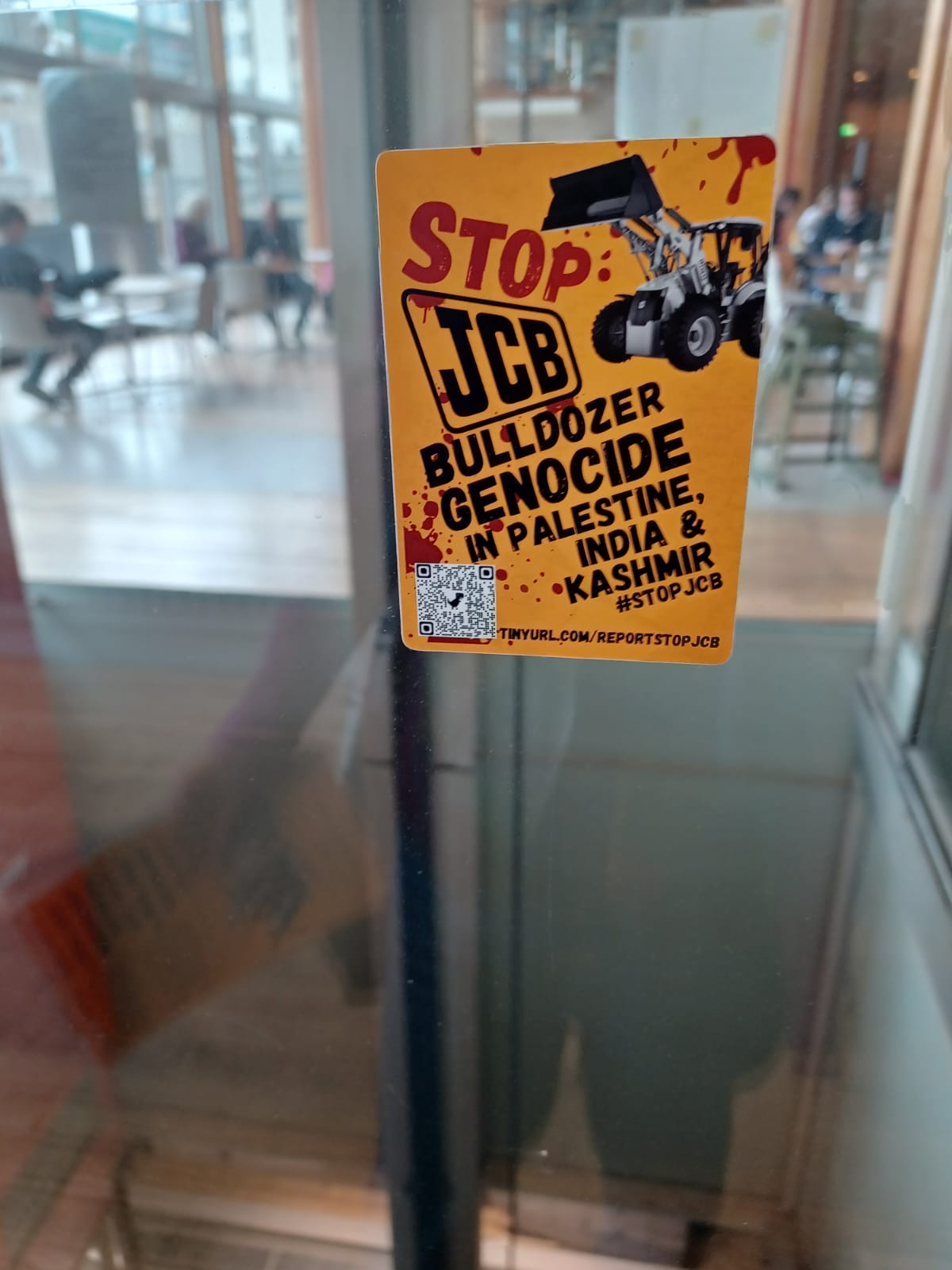 Sticker on a glass panel in a building where a the JCB sponsored lift is which reads: "Stop JCB bulldozer genocide in Palestine, India & Kashmir. #STOPJCB