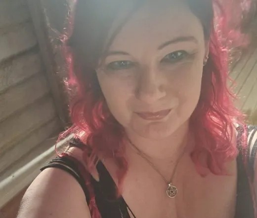 Jayne Campbell. Woman with reddish-pink hair down to her shoulders, a black top, and silver pentagram necklace.