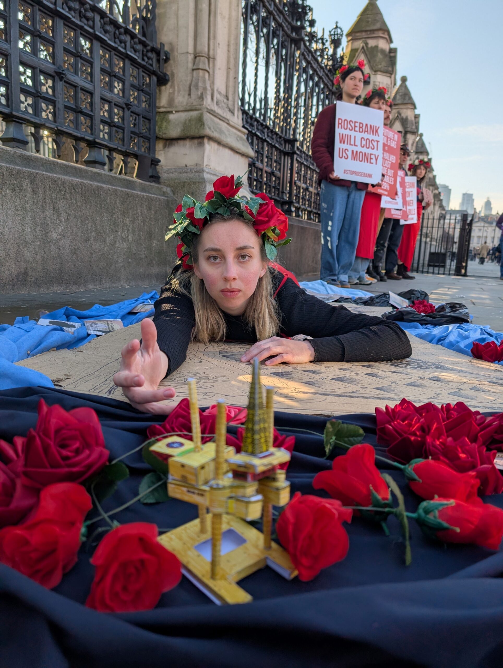 Campaigner lays on a blue sheet on the pavement reaching out her hand to a 3D model of an oil rig surrounded by roses. 