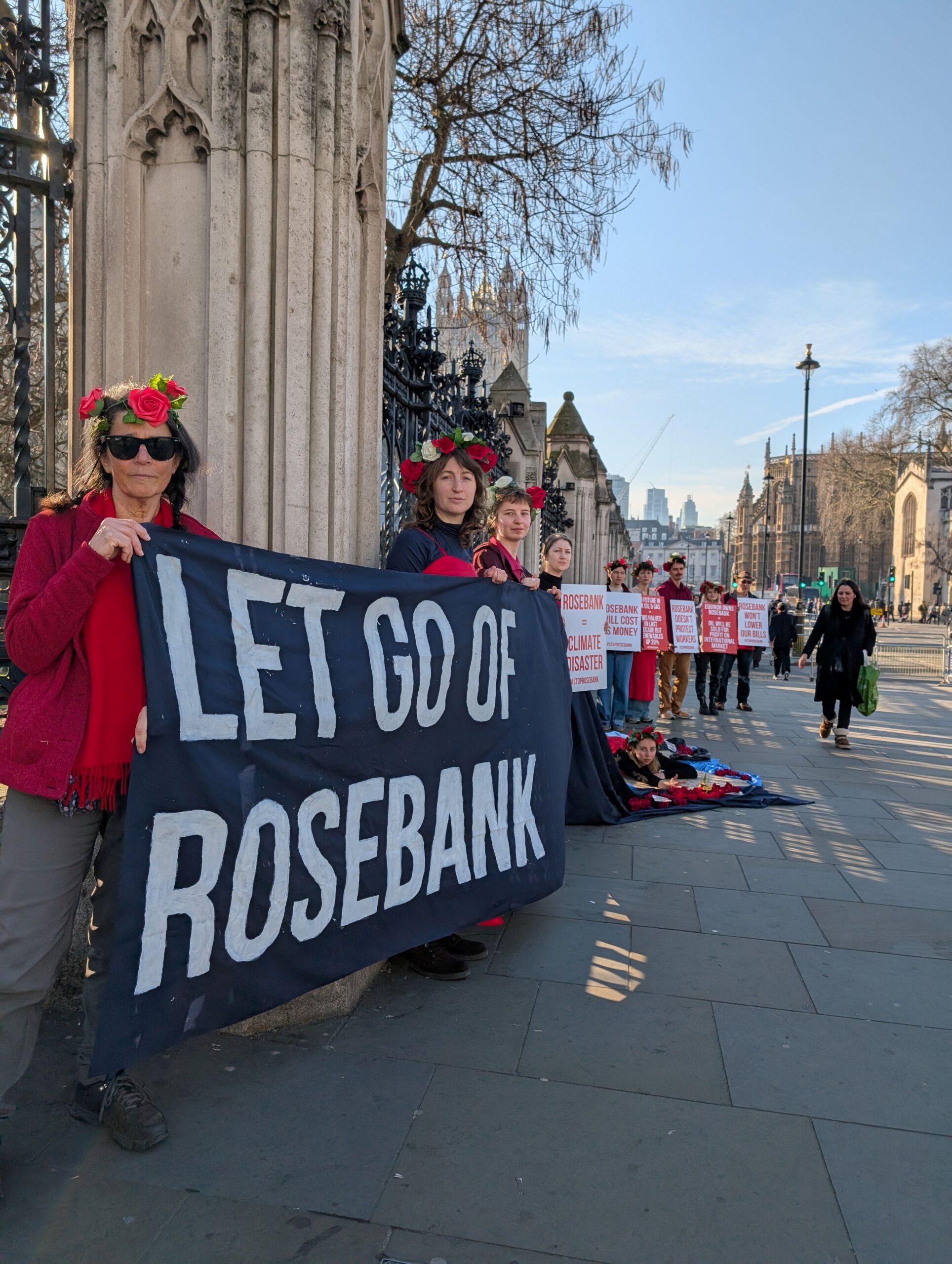 Protesters in red rose crowns hold a banner that reads "Let go of Rosebank". 