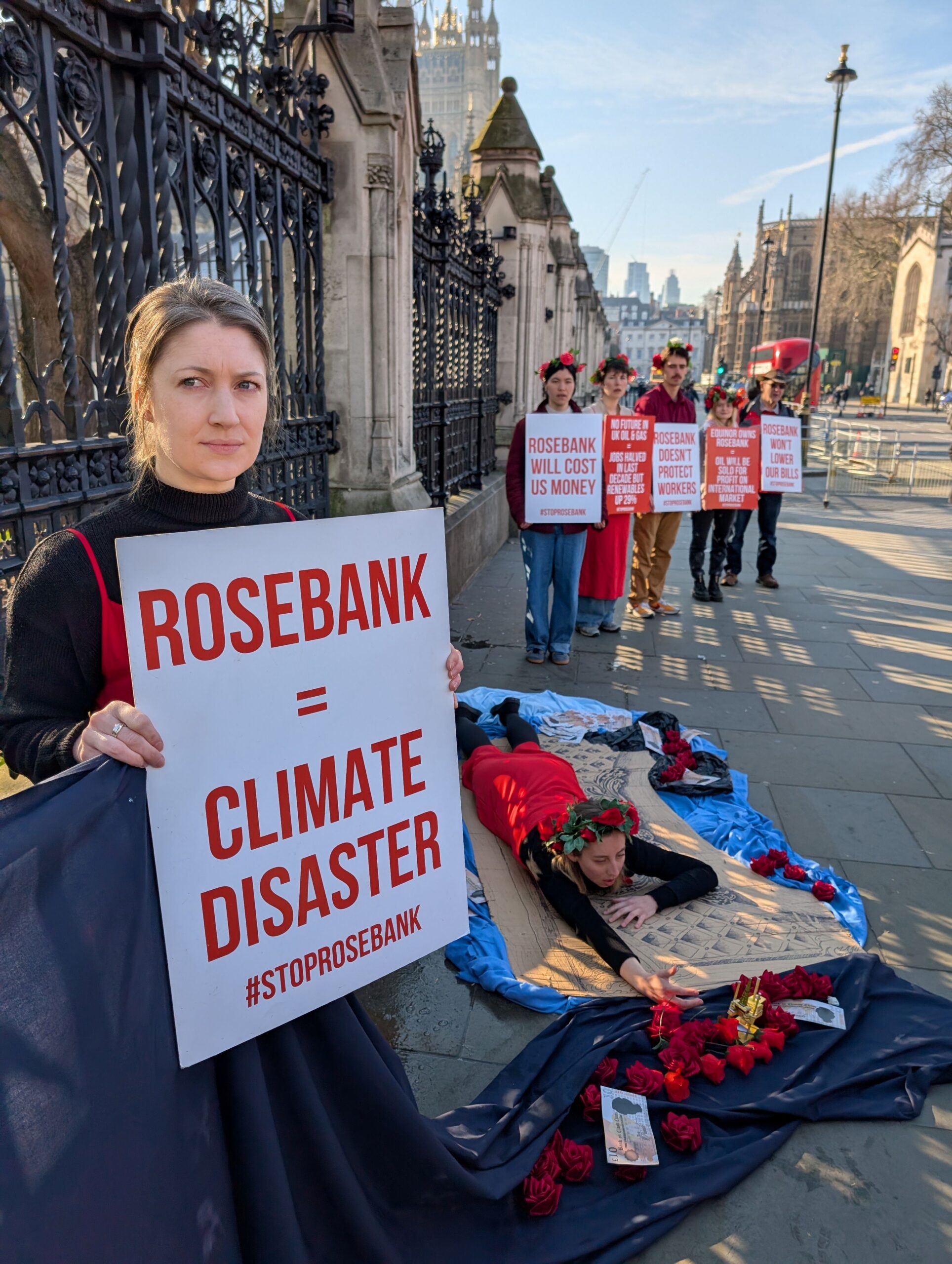 Protester lays on the pavement re-enacting the Titanic scene where Rose lets go of Jack's hand. She reaches out to a 3D smallscale model of an oil rig surrounded by roses. Campaigner in the foreground holds a placard that reads: "Rosebank = Climate disaster #STOPROSEBANK" Other protesters hold placards in the distance. Closest reads: "Rosebank will cost us money." 