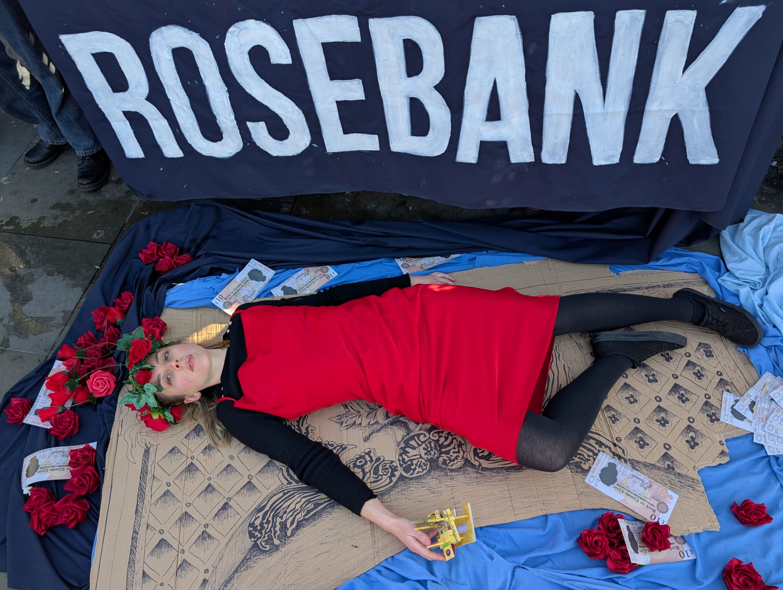 Protester lays on the pavement re-enacting the Titanic scene on the piece of ship she's using as a raft. She holds a 3D smallscale model of an oil rig surrounded by roses. 