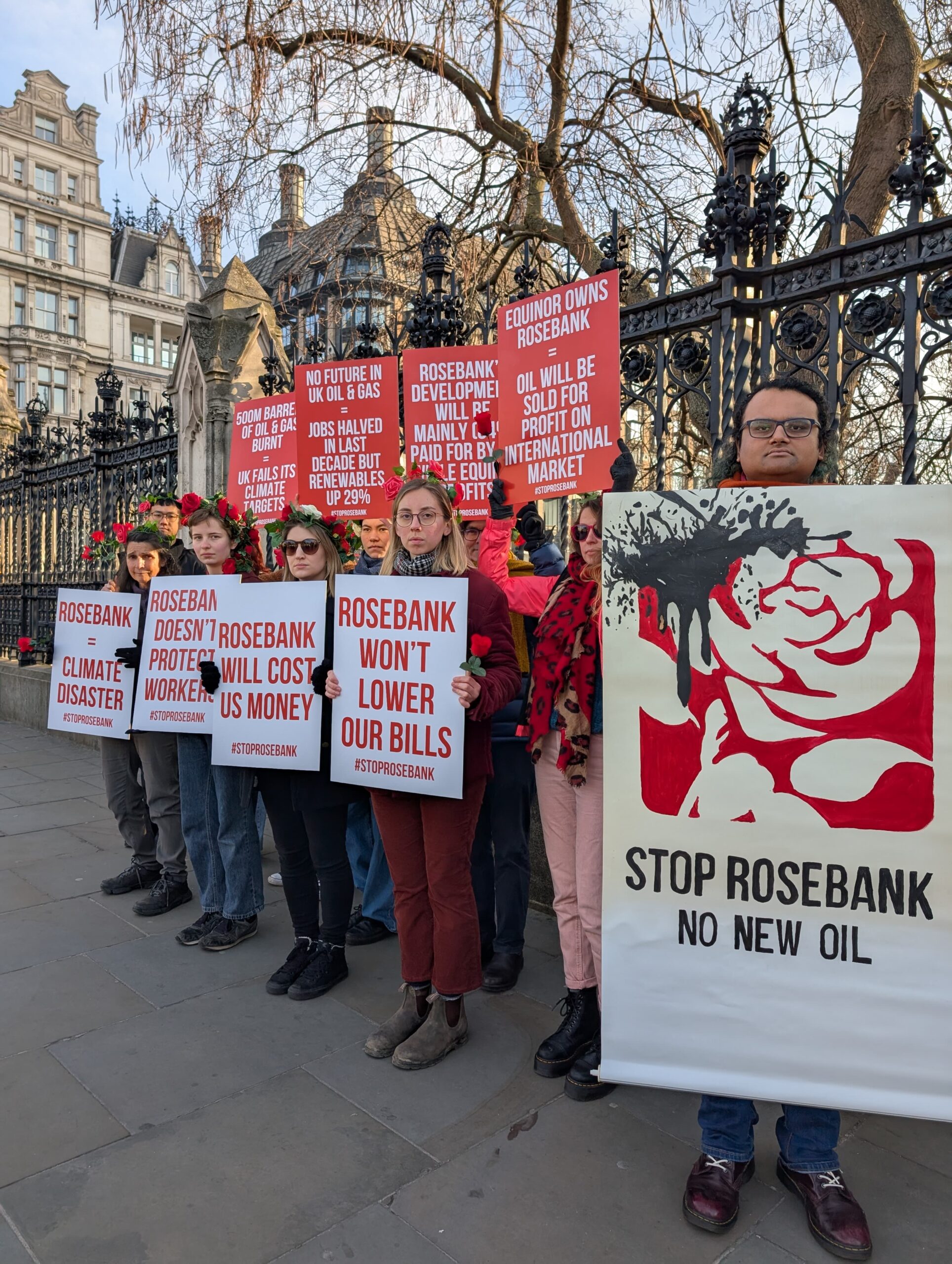 Campaigners adorned in roses hold placards with Parliament in the background. These read from left to right, top to bottom: "500m barrels of oil & gad burnt = UK fails its climate targets", "No future in UK oil & gas = jobs halved in last decade but renewables up 29%", "Rosebank's development will be mainly (84%) paid for by us while Equinor profits", "Equinor owns Rosebank = oil will be sold for profit on international market", "Rosebank = climate disaster", "Rosebank doesn't protect workers", "Rosebank will cost us money", and "Rosebank won't lower our bills". The final poster to the right displays the Labour Party rose logo dripping in oil with the caption "Stop Rosebank - no new oil". 