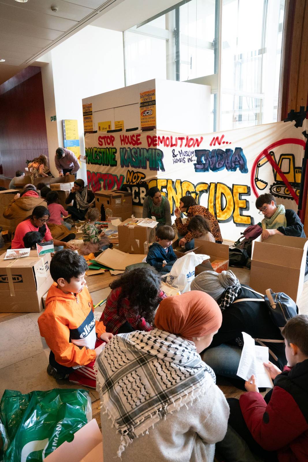 Parents and children sit amid boxes and craft supplies in a room with a bold banner reading: "JCB - stop house demolition in Palestine, Kashmir & Muslim homes in India. Poetry not JCB genocide." Illustration of a JCB digger with a red stop sign crossed over it. 